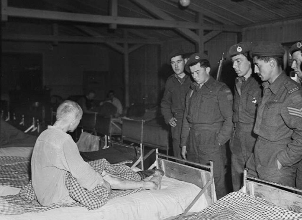 Ein abgemagerter Häftling sitzt auf einem Bett, vor ihm stehen amerikanische Soldaten.